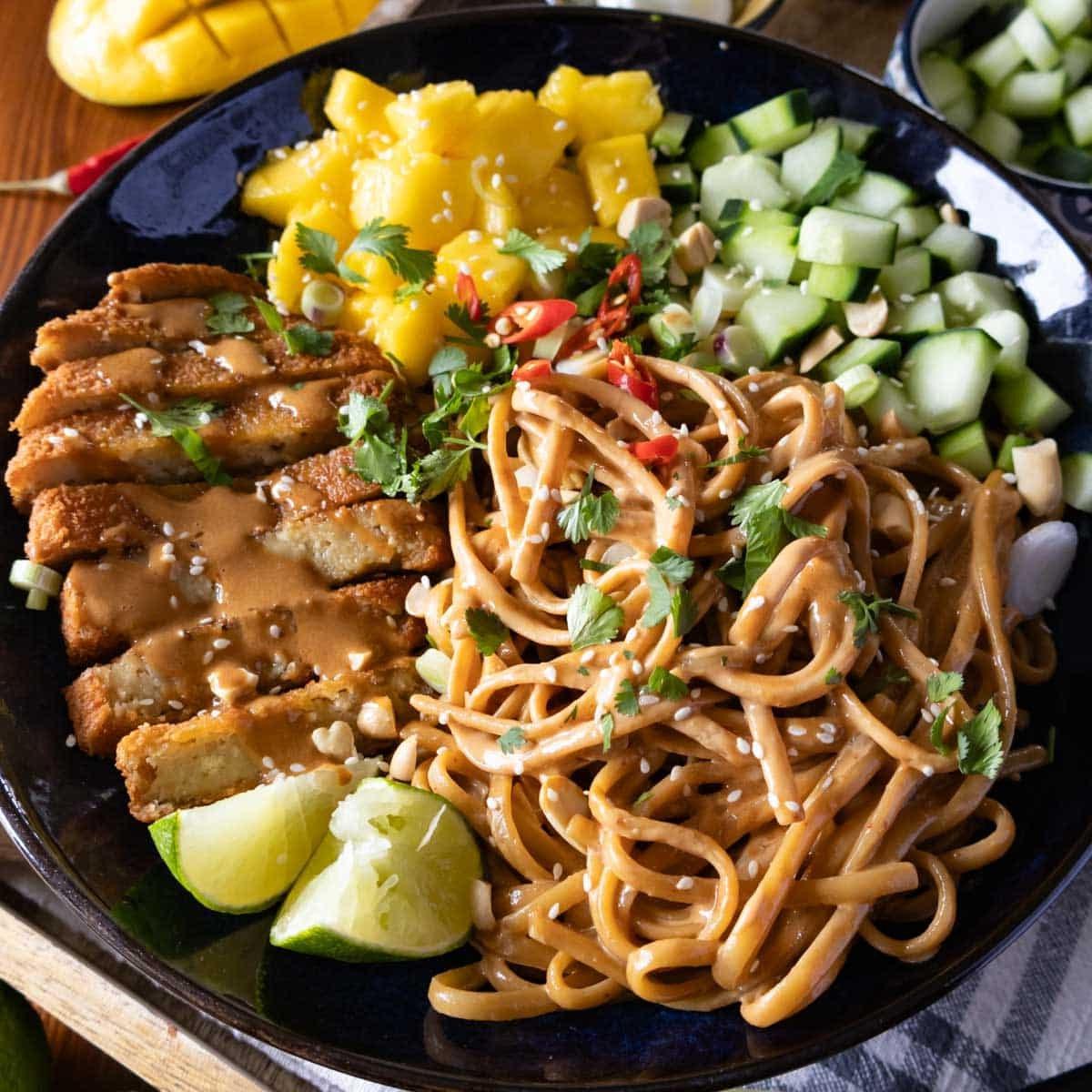 Sesamnudeln in einer dunklen Schüssel, garniert mit Mango, Gurke und Limette.