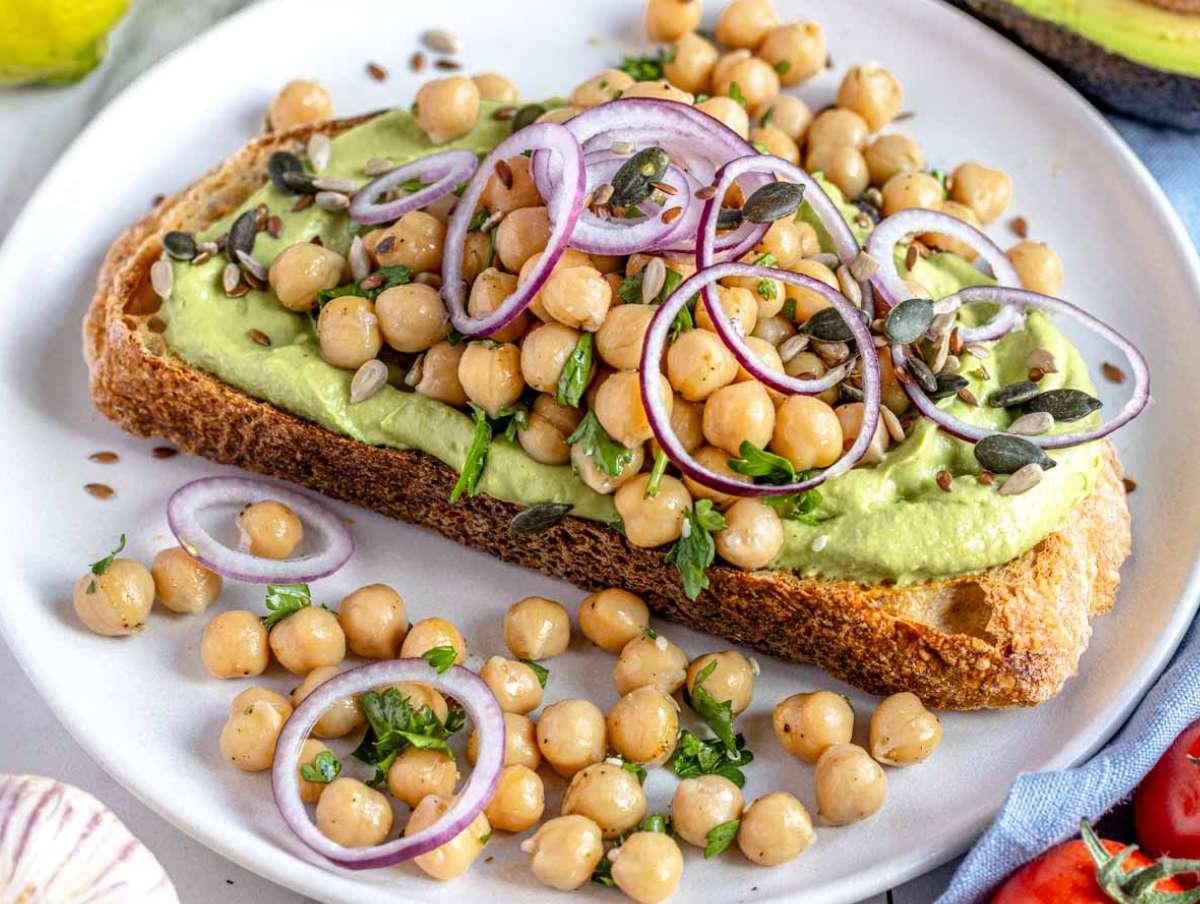 Avocado-Aufstrich auf einer Scheibe geröstetem Brot, belegt mit marinierten Kichererbsen und Kräutern.