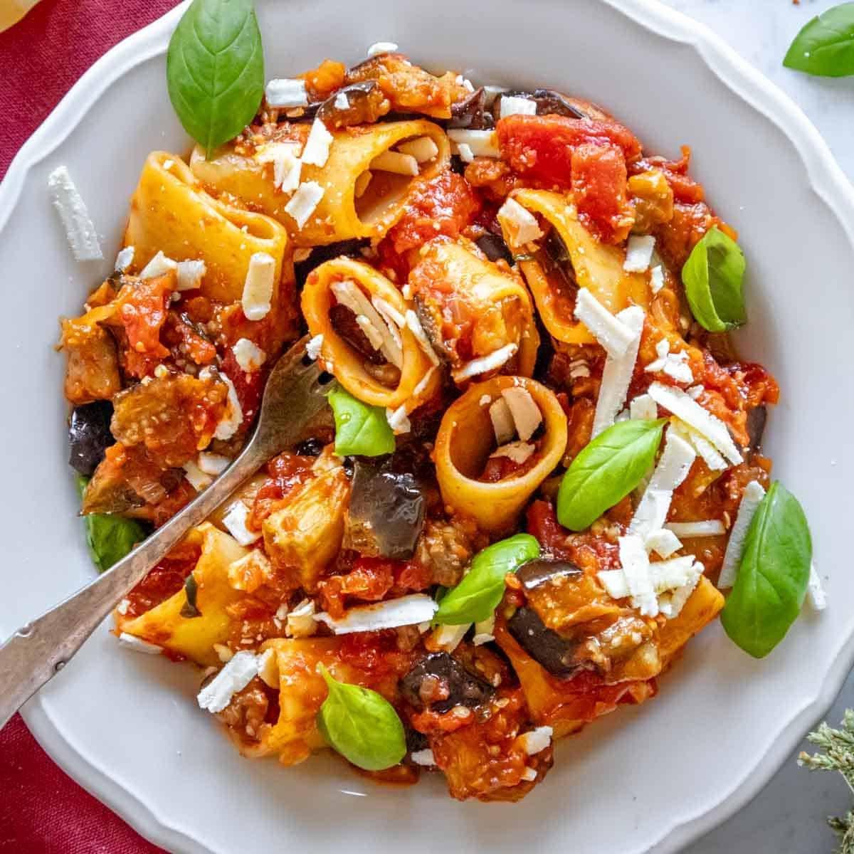 Pasta mit Aubergine auf einem weißen Teller mit frischem Basilikum und einer Gabel.