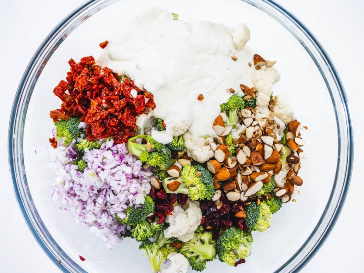 broccoli cauliflower, dressing, nuts, and red onions in a glass bowl