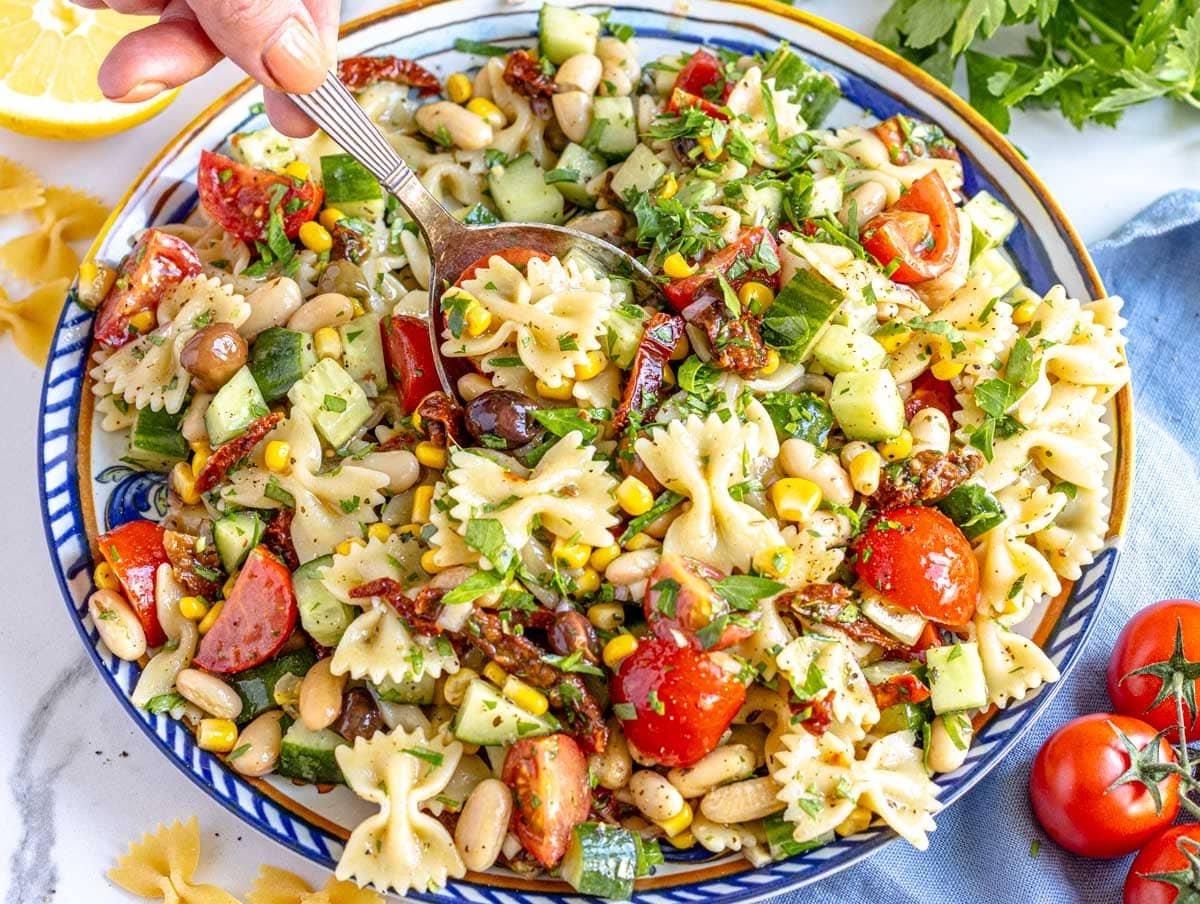 Veganer Nudelsalat auf einem blauen Teller mit einer Hand, die einen silbernen Löffel hält.